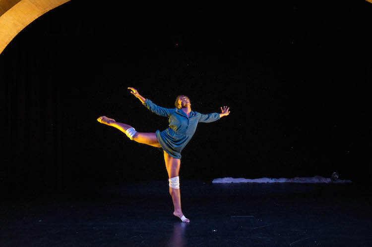 Kayla Farrish in a deep blue longsleeved mini-dress spreads her arms wide like a bird as she floats in a high arabesque position her knees are covered with white knee pads.